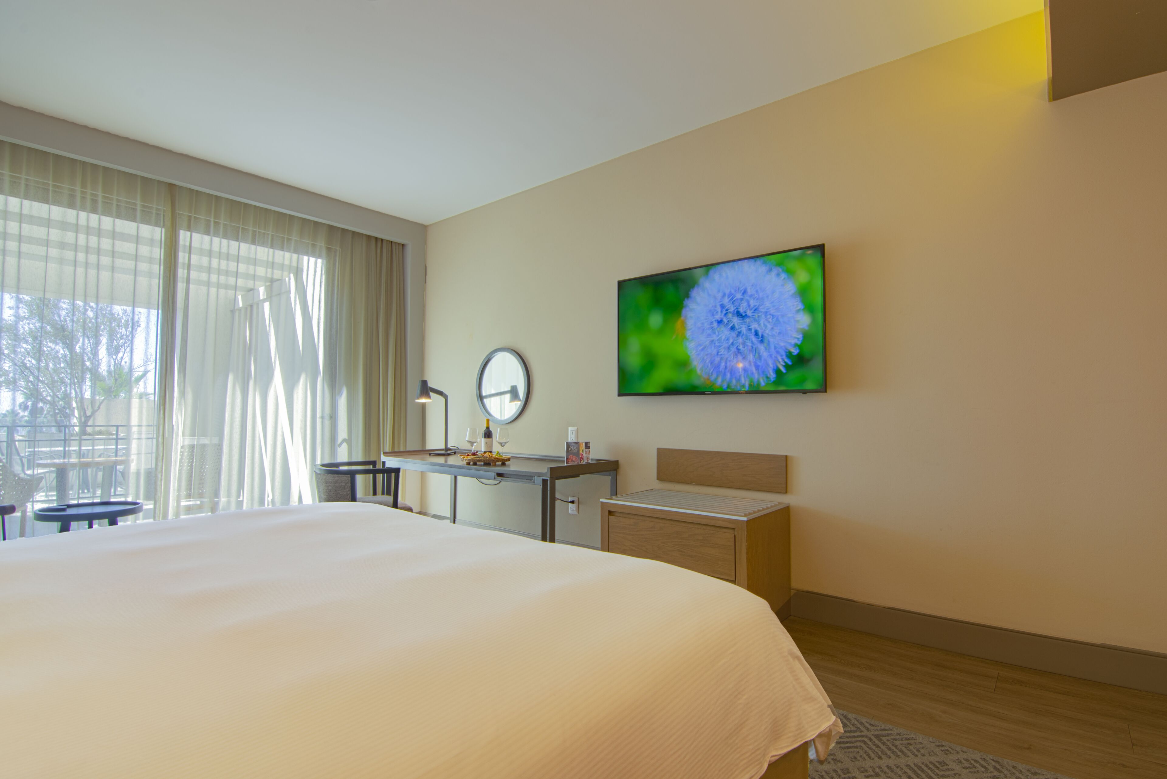 A hotel room with a large bed, a desk with a lamp, a TV on the wall, and a window with curtains.