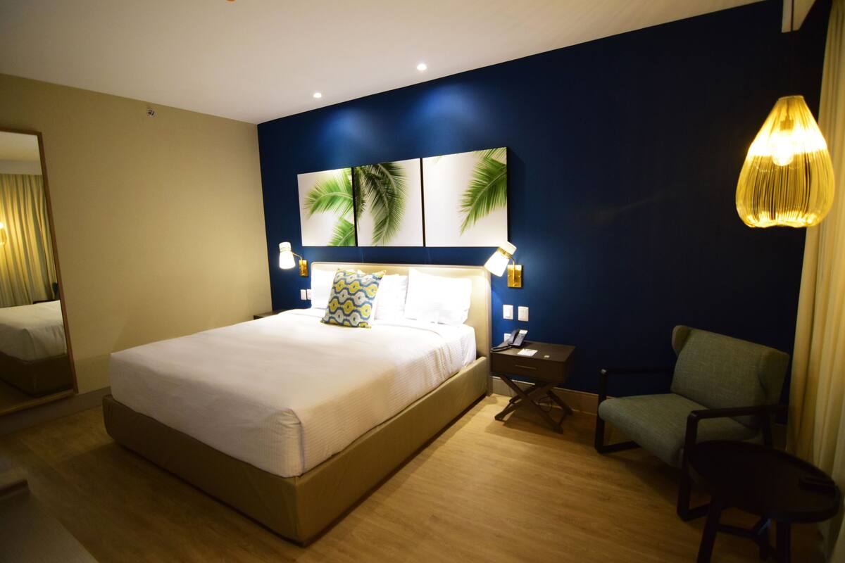 A hotel room with a large bed, a chair, a nightstand, and a wall with palm leaf artwork.