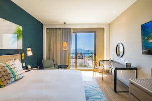 Habitación de hotel con cama, escritorio, silla, balcón con vistas y un cuadro en la pared.