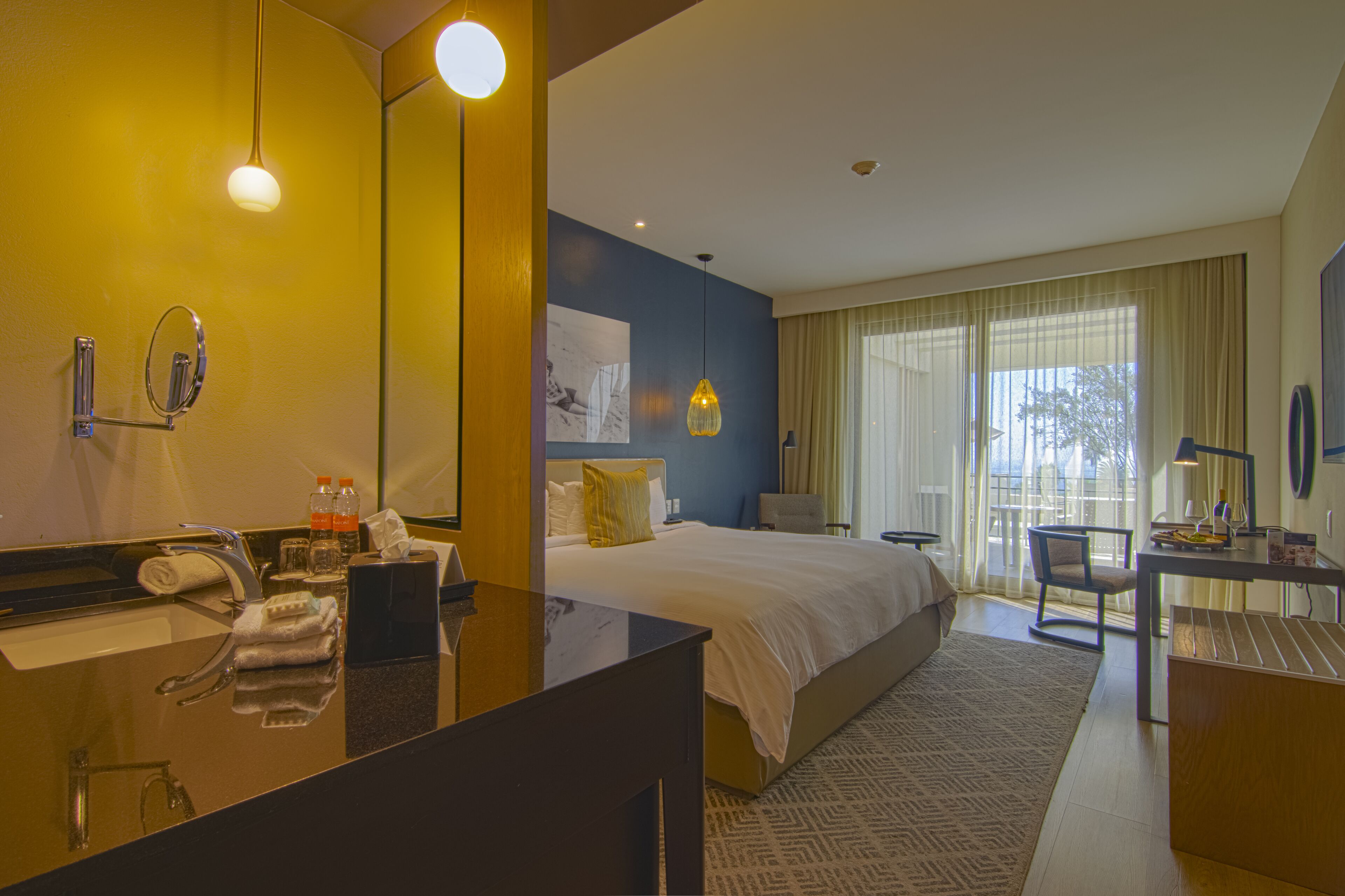 A hotel room with a large bed, a desk, a chair, a bathroom with a mirror, and a window with curtains.