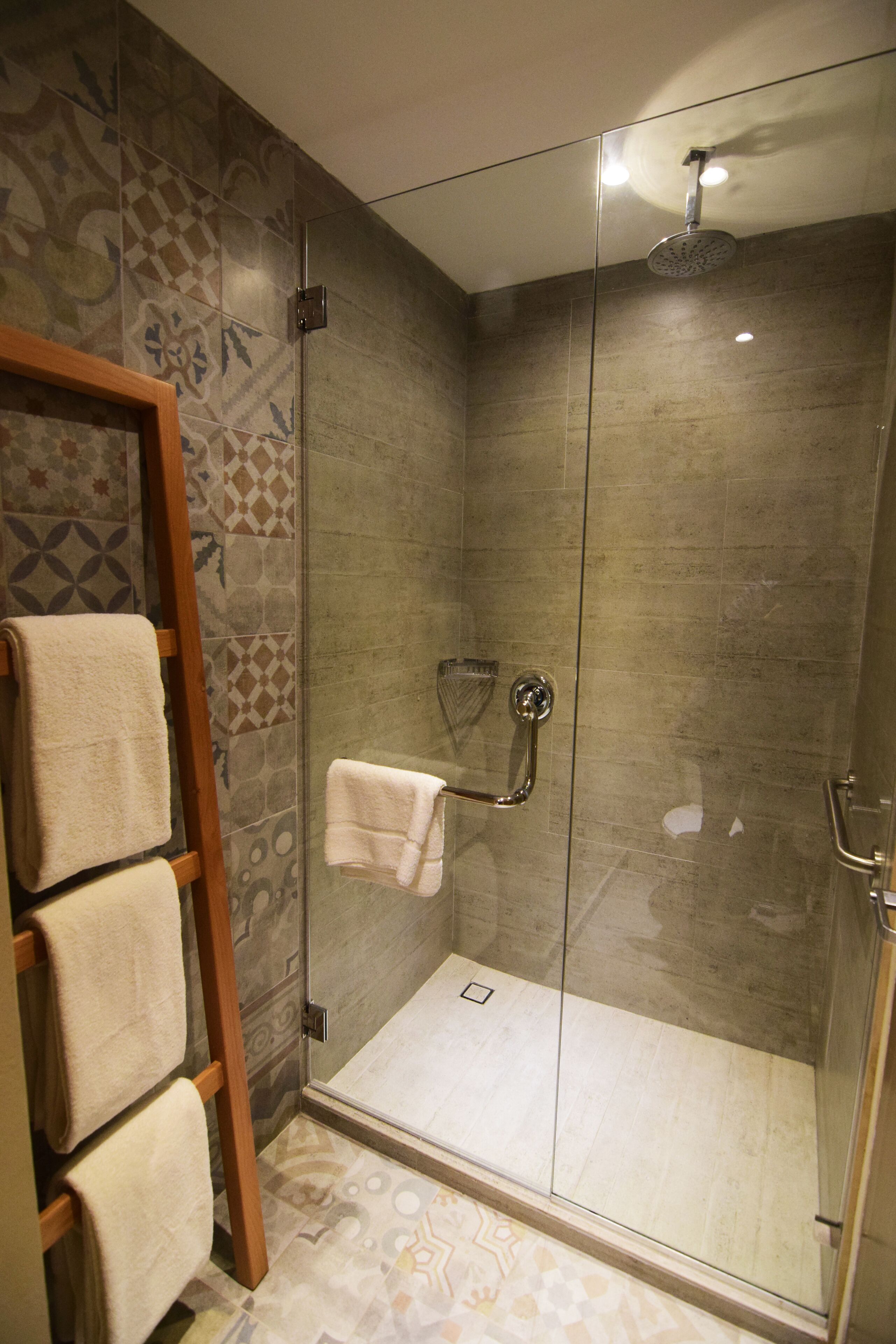 A glass-enclosed shower with a handheld showerhead, a towel rack with multiple towels, and patterned floor tiles.