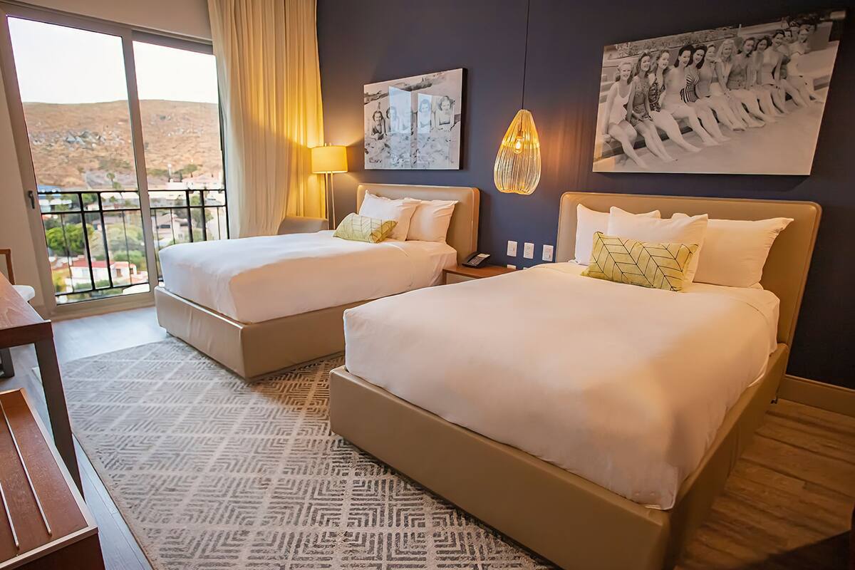 A hotel room with two beds, a desk, a chair, a patterned rug, and a balcony with a view.