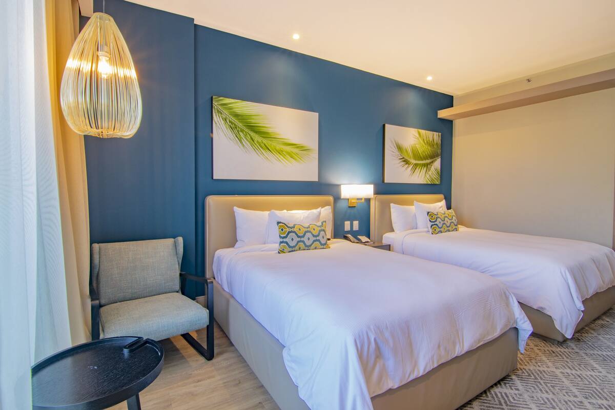 A hotel room with two beds, a chair, a small table, and wall art.