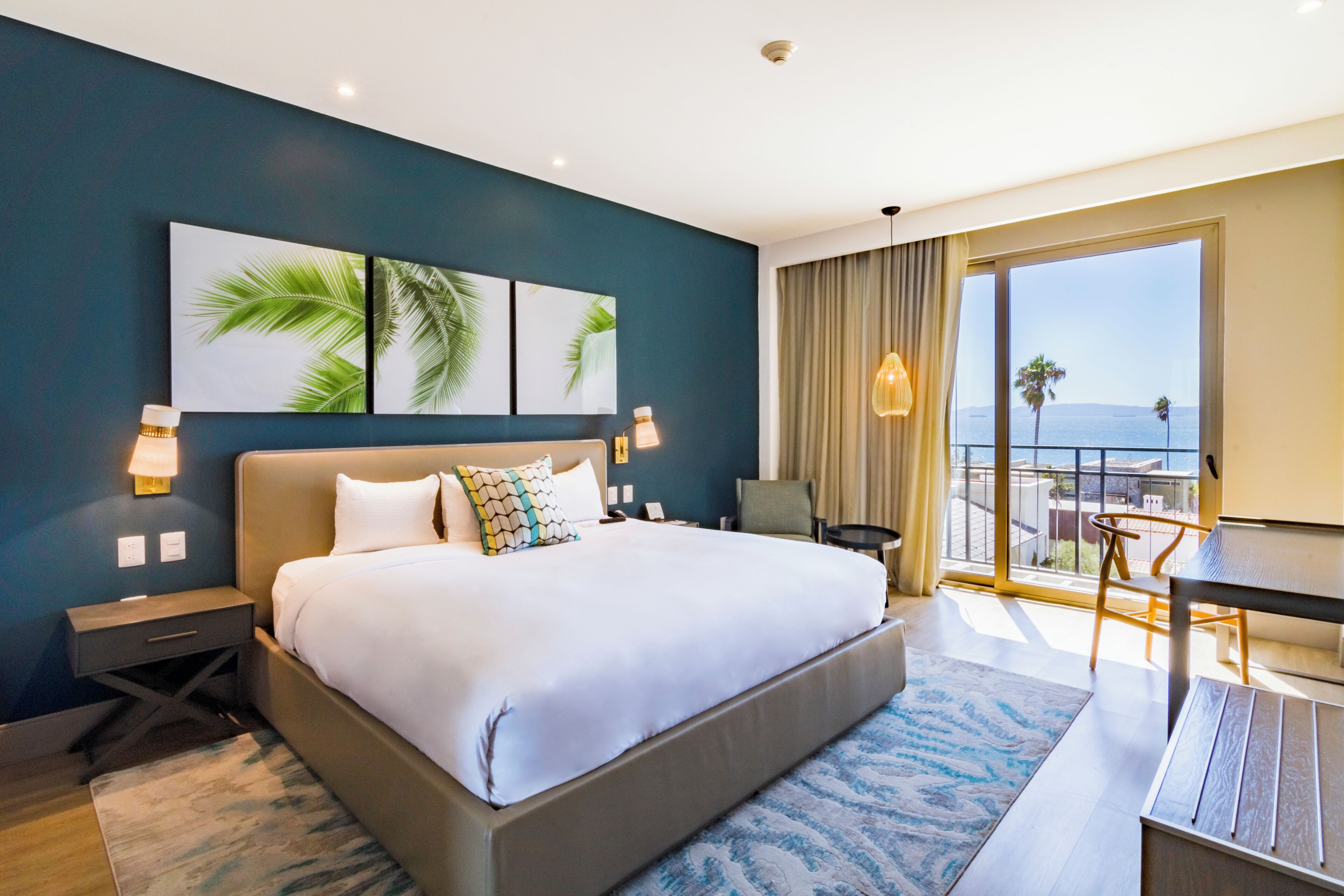 Una habitación de hotel con una cama grande, un escritorio, una silla, vista al mar y una obra de arte con hojas de palma en la pared.