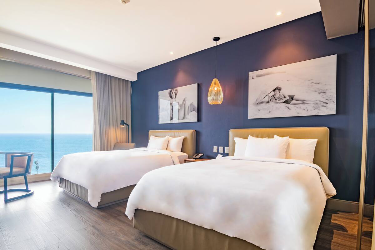 A hotel room with two beds, a large window with a view of the sea, and artwork on the walls.