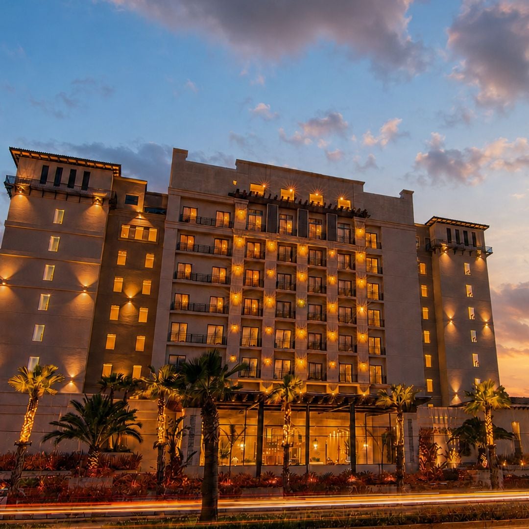 A multi-story hotel with a symmetrical facade, illuminated windows, and a landscaped front with palm trees.
