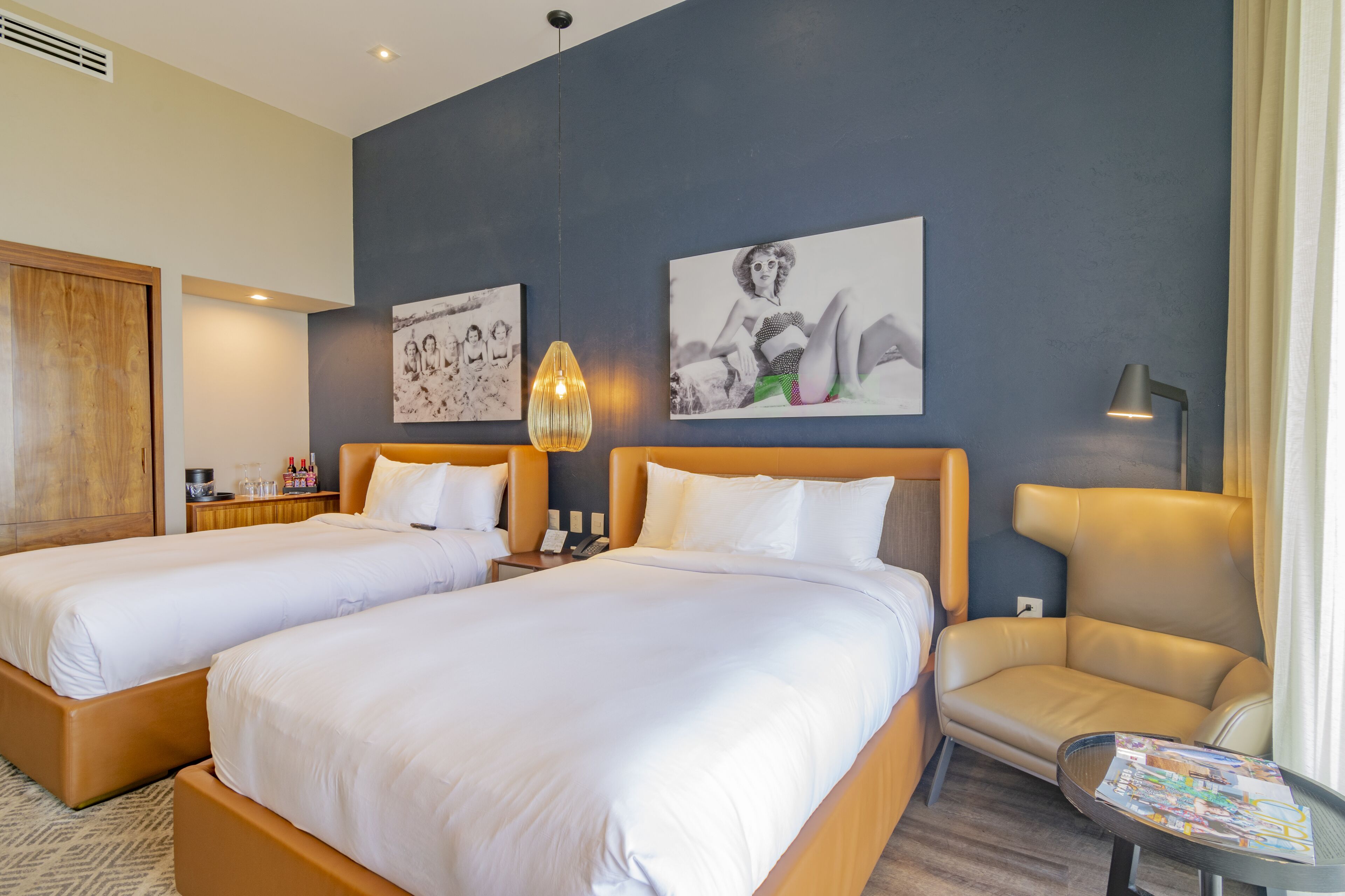 A hotel room with two beds, a chair, a table, and artwork on the wall.