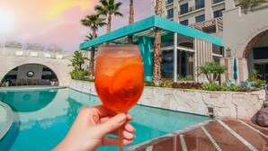 Eine Hand hält ein Glas mit rotem Getränk, Poolblick auf ein Hotelgebäude und Palmen.