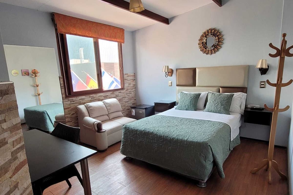 Habitación de hotel con cama, sofá, escritorio, perchero y ventana con banderas.
