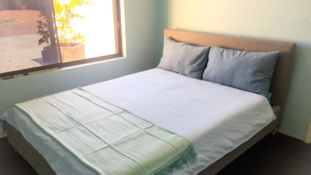 A bed with a white sheet and a green throw blanket, a wooden headboard, and a window with a view of a street and a plant outside. - Caversham B&B (Beechboro)