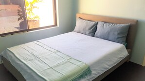 A bed with a white sheet and a green throw blanket, a wooden headboard, and a window with a view of a street and a plant outside. - Caversham B&B (Beechboro)