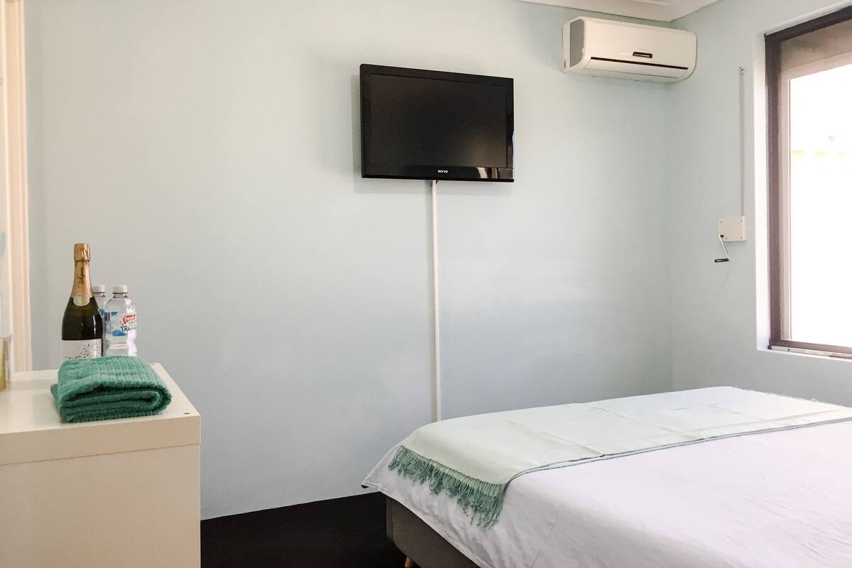 Une chambre d’hôtel avec un lit, un téléviseur à écran plat fixé au mur, un appareil de climatisation, une fenêtre et une petite table sur laquelle se trouvent une bouteille de champagne et une serviette.