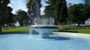 Une fontaine avec des jets d’eau dans un bassin circulaire, entourée d’une allée pavée et de verdure.