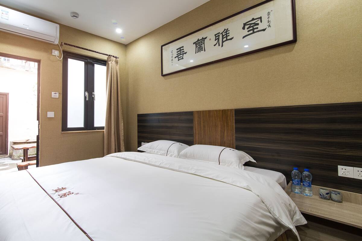 Une chambre d’hôtel comprenant un lit, une tête de lit, une fenêtre avec des rideaux, un appareil de climatisation et un panneau fixé au mur.