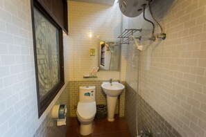 A bathroom with a toilet, sink, and shower. There is a window with blinds, a towel dispenser, and a trash can.