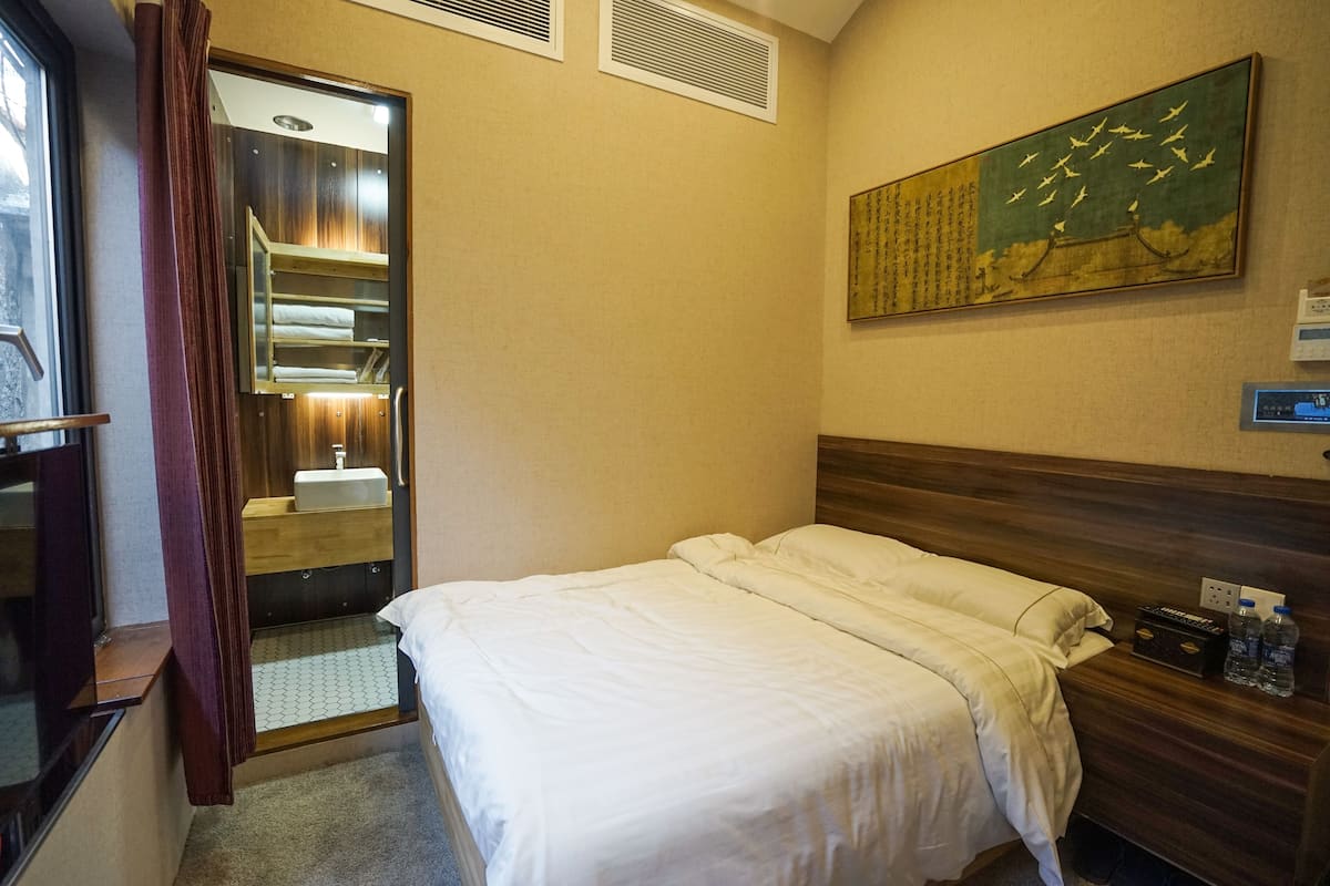 Une chambre d’hôtel avec un lit, une table de chevet avec une radio, un tableau au mur et une salle de bain avec un miroir et un lavabo.