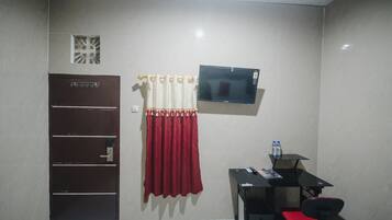 A small room with a door, a TV mounted on the wall, a desk with a chair, and a red curtain.