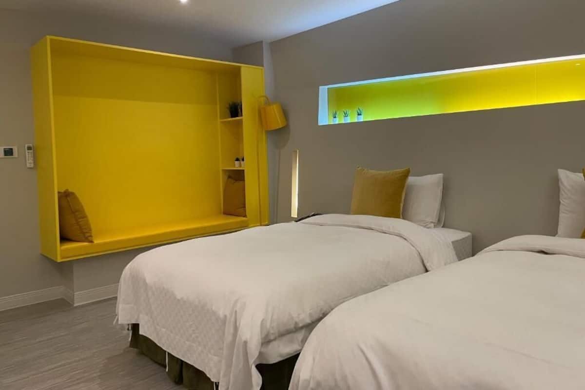 A modern hotel room with two beds, a yellow built-in bench, and a shelf with plants.
