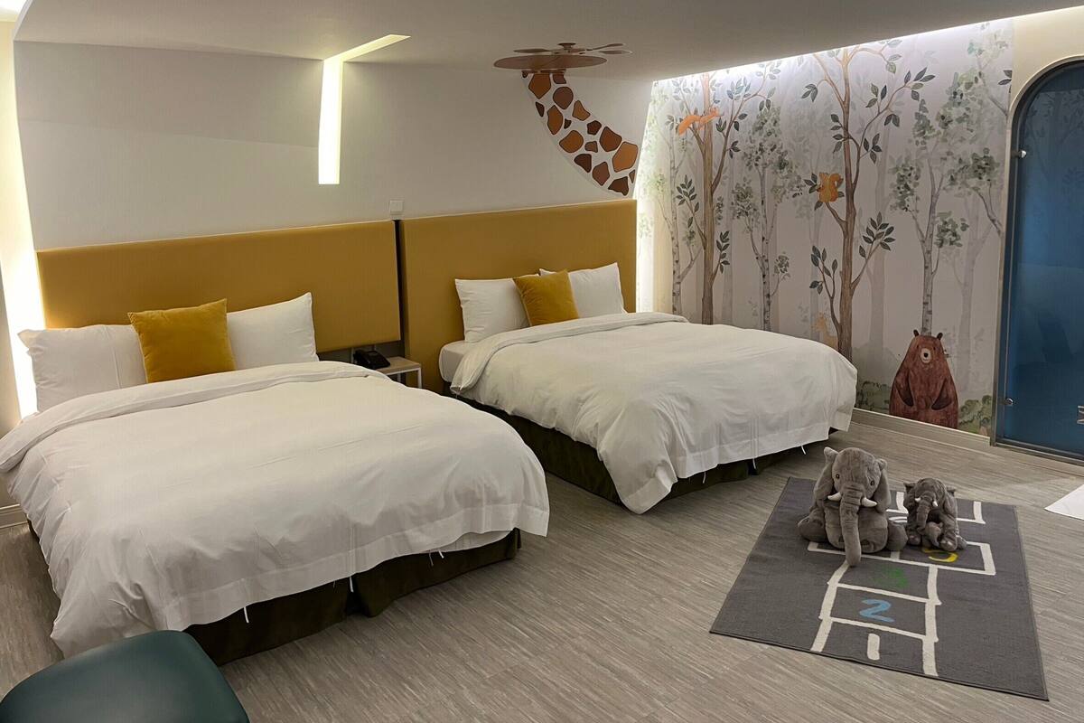 A hotel room with two beds, a giraffe mural, and a bear and elephant play mat.
