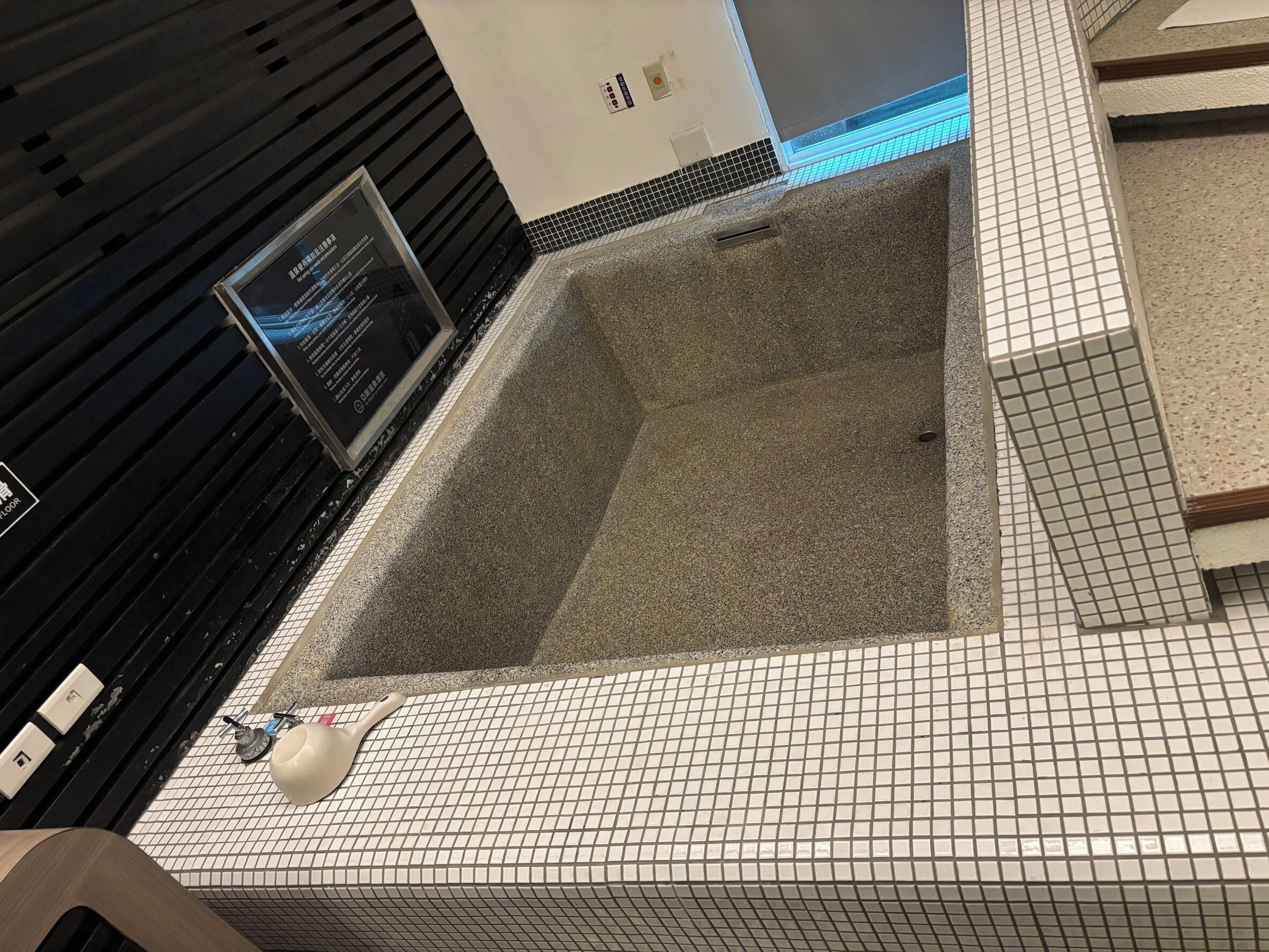 A tiled floor with a rectangular area that appears to be a hot tub or a small pool. There is a sign with text on the wall to the left, and a light fixture above the tiled area.