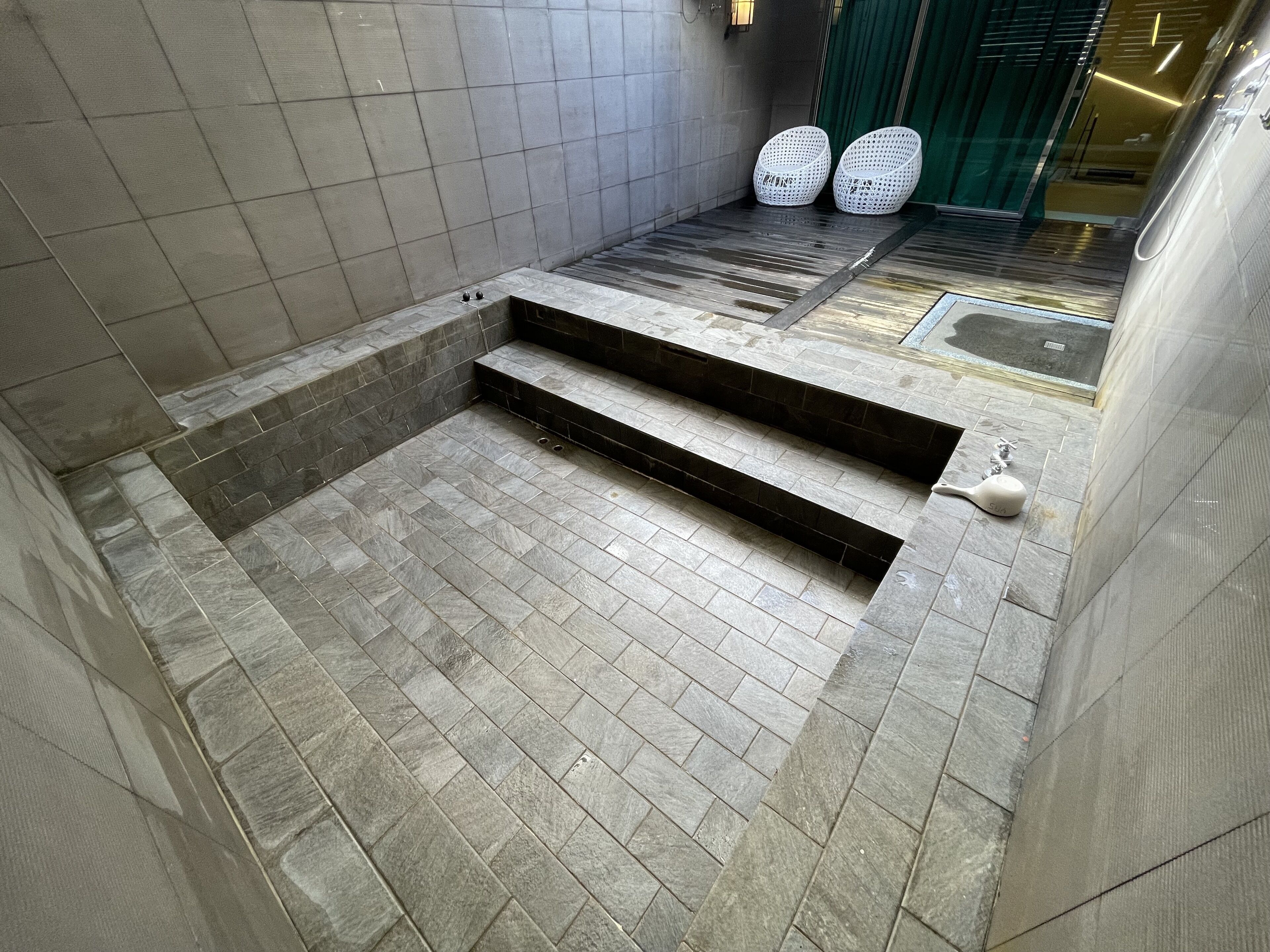Un baño con paredes y suelo de baldosas grises, un lavamanos pequeño y dos sillas de mimbre blancas.