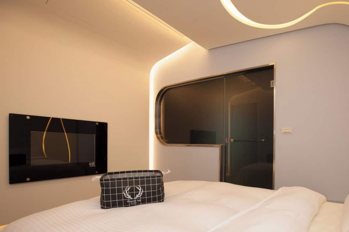 A modern hotel room with a curved ceiling, a large window, a flat-screen TV, and a neatly made bed.