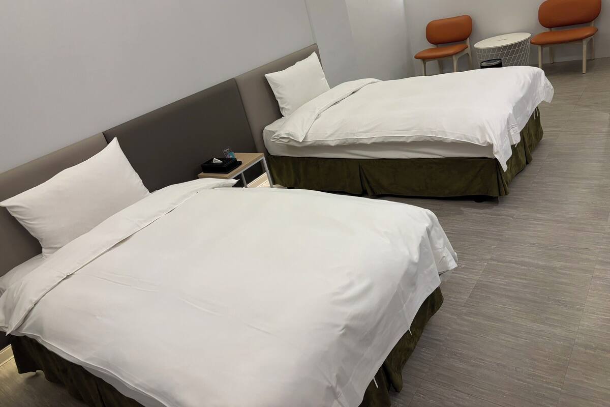 A hotel room with two beds, a small table with a chair, and a window.
