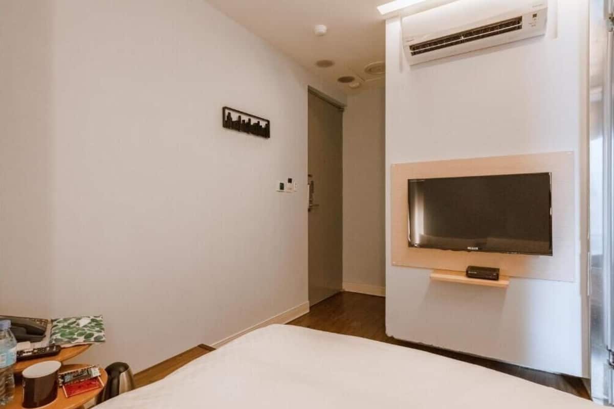 A hotel room with a bed, a mounted TV, an air conditioning unit, and a small table with a remote control and a water bottle.
