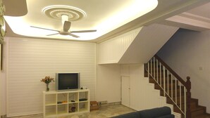 A living room with a TV on a white stand, a ceiling fan, a staircase, and a sofa. - Bukit Tinggi Klang - Cozy Home (Klang)