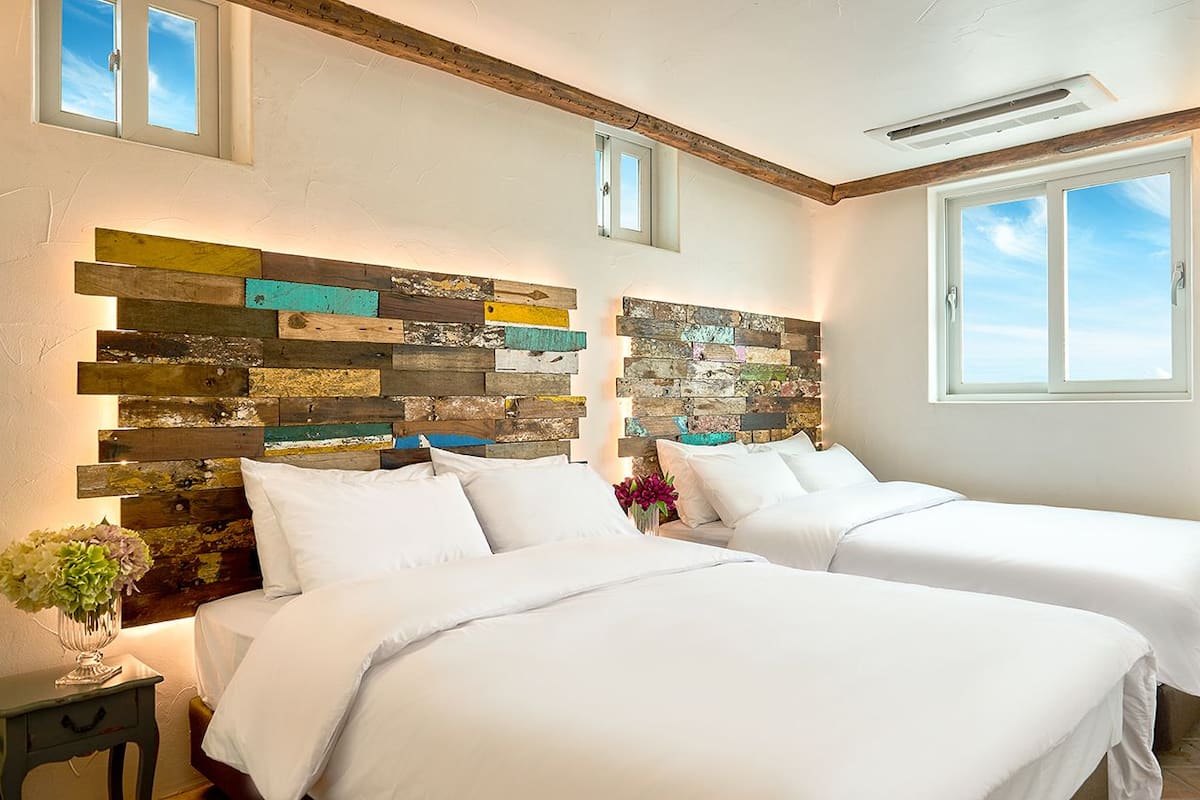 A hotel room with two beds, a rustic wooden headboard, a small table with a vase of flowers, and a large window with a view.
