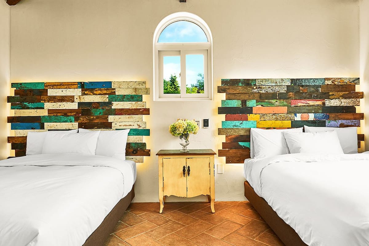 A hotel room with two beds, a small wooden nightstand with a vase of flowers, and a rustic wall panel design.