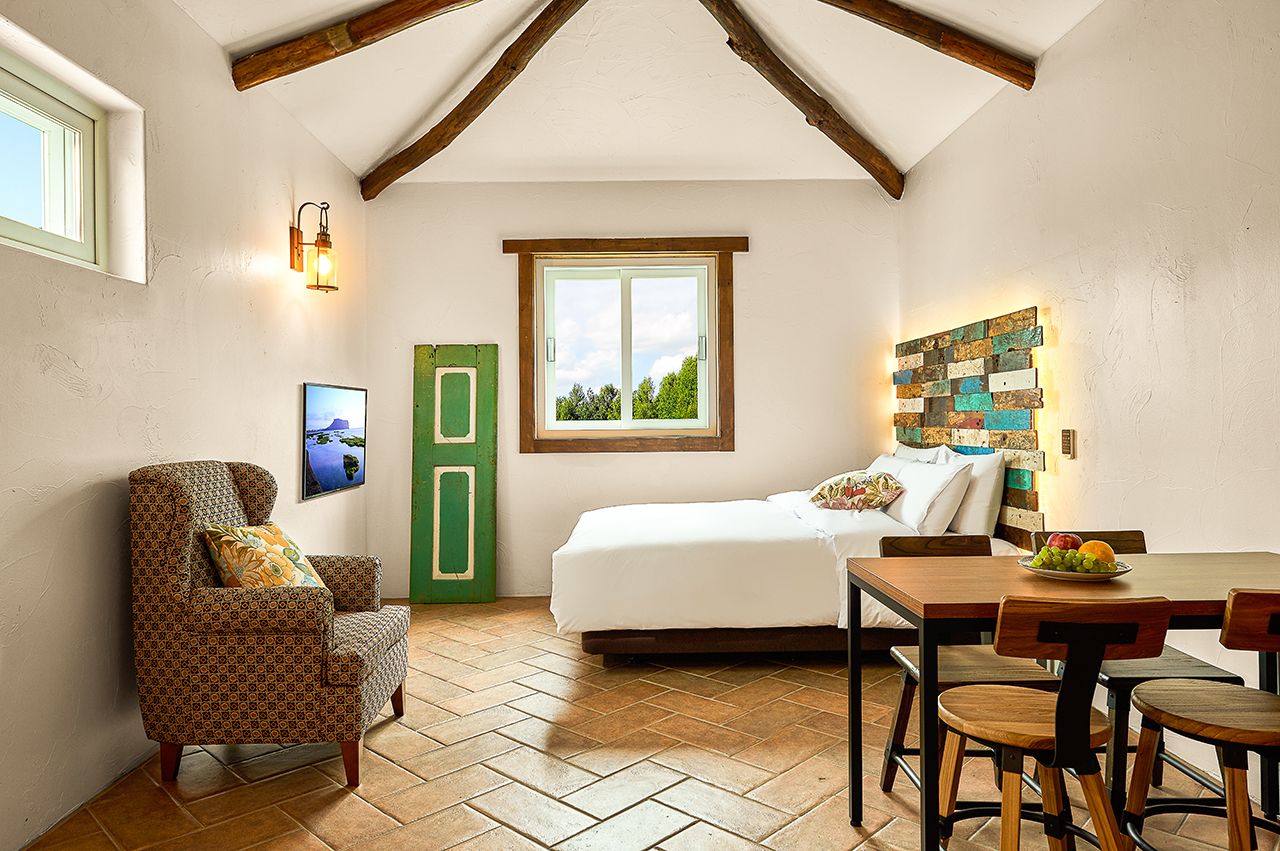Quarto aconchegante com cama, mesa de jantar e cadeiras, porta verde e luminária fixada na parede.
