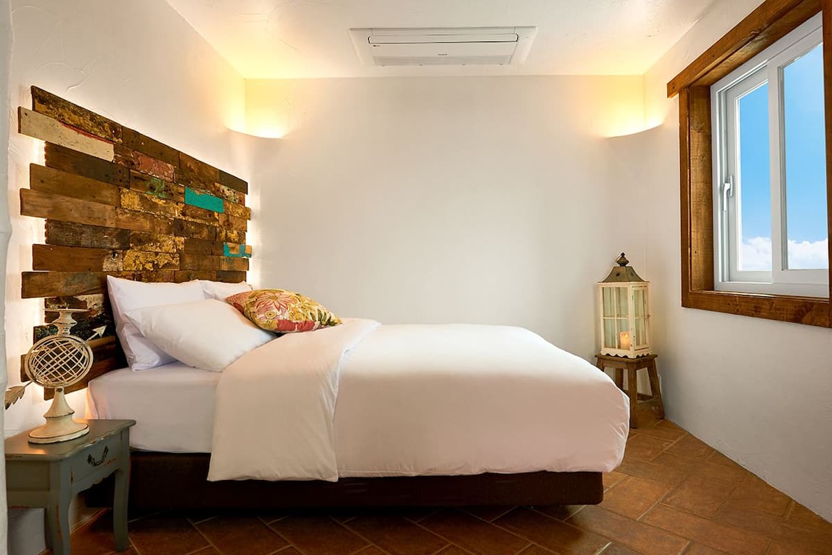 A bedroom with a wooden headboard, a bed with white linens, a nightstand with a lamp, and a window with a view of the sky.