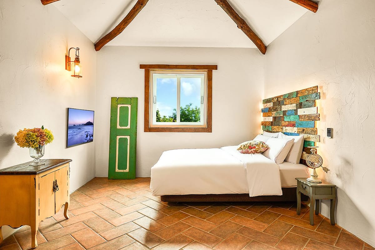A bedroom with a bed, a green door, a window, a wall-mounted light fixture, and a decorative wall piece.