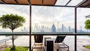 A modern outdoor seating area with a glass enclosure, a wooden deck, and a view of a city skyline. - Wong's Homestay at Regalia Residence (Kuala Lumpur)