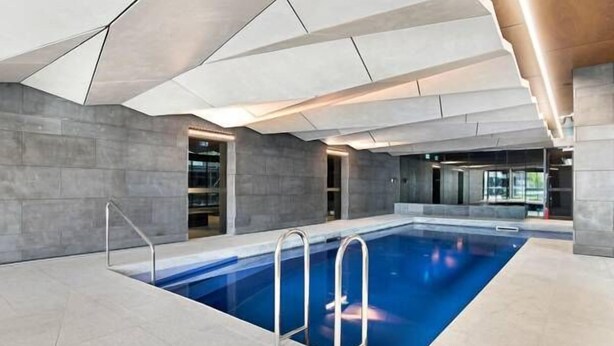 A modern indoor swimming pool with a geometric ceiling design, clear pool area, and metal ladders. - Southern Cross CBD Apartment (Melbourne)