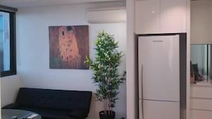 A modern living room with a black sofa, a dining table, a potted plant, and a white refrigerator. - Southern Cross CBD Apartment (Melbourne)