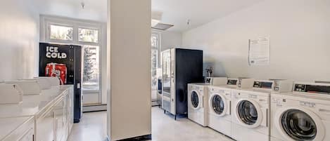 A laundry room with multiple washing machines, a refrigerator, and a window with a view of trees.