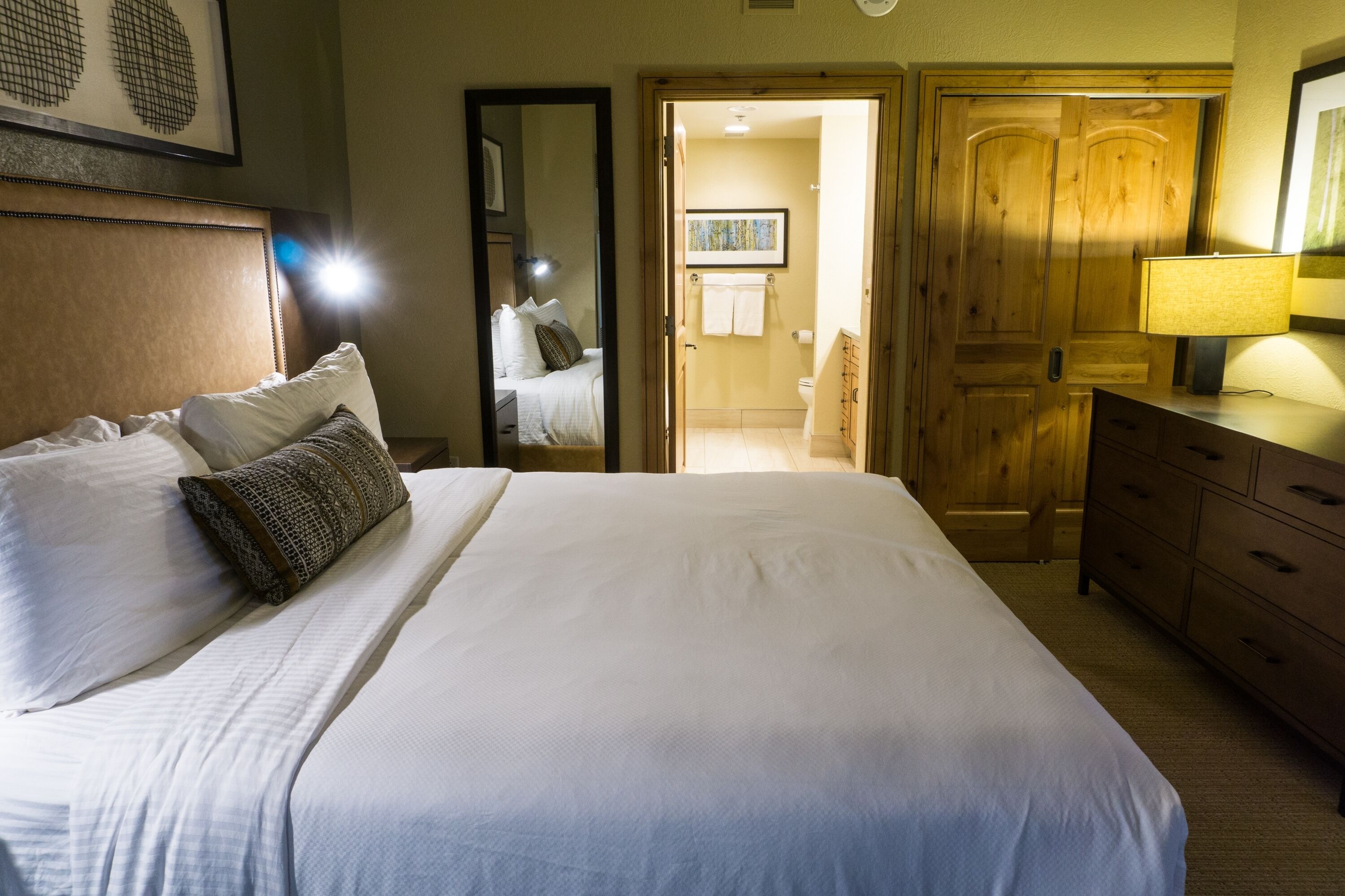 A neatly made bed with white linens and a patterned pillow, a wooden dresser, and a wooden door leading to a bathroom.