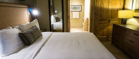 A neatly made bed with white linens and a patterned pillow, a wooden dresser, and a wooden door leading to a bathroom.