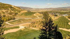 A golf course with a winding path, a large tree in the foreground, and a distant view of a town with houses and a hill in the background.