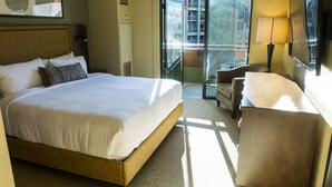 Ett hotellrum med en stor säng, ett skrivbord, en stol, en balkong med utsikt över byggnader och en tv som är monterad på väggen.