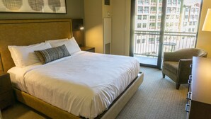 A hotel room with a large bed, a nightstand, a chair, a desk, and a window with a view of buildings.