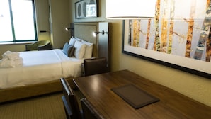 Quarto de hotel com cama, escrivaninha, cadeira, luminária e uma grande janela.