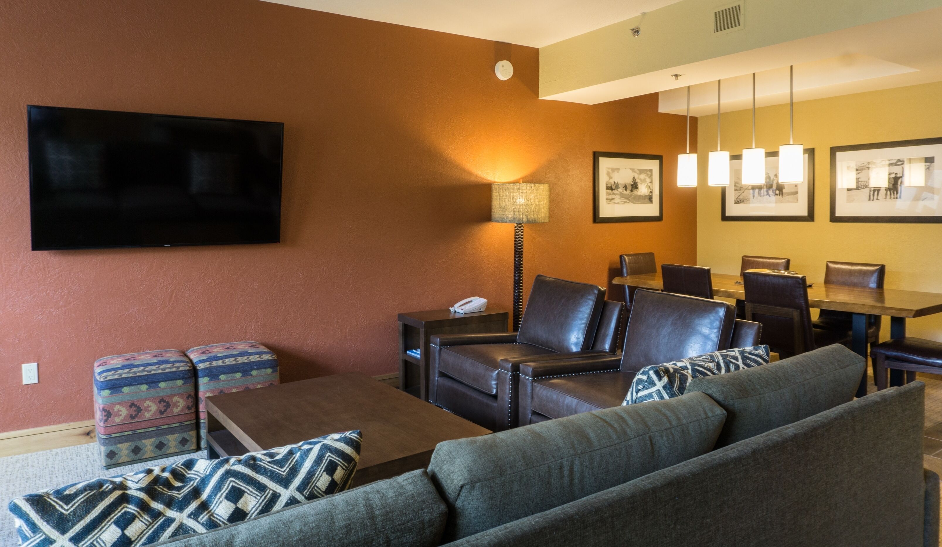 A modern living room with a flat-screen TV, leather sofa, patterned ottoman, and a dining area with a table and chairs.