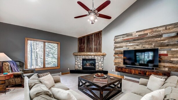 A modern living room with a stone fireplace, a large flat-screen TV mounted on a wooden wall, and a ceiling fan. - Streamside 333B (Frisco)