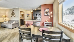 A cozy living room with a gray sofa, a wooden dining table with four chairs, a kitchen area with a microwave, and a large window with a snowy view. - Forest Park F (Frisco)