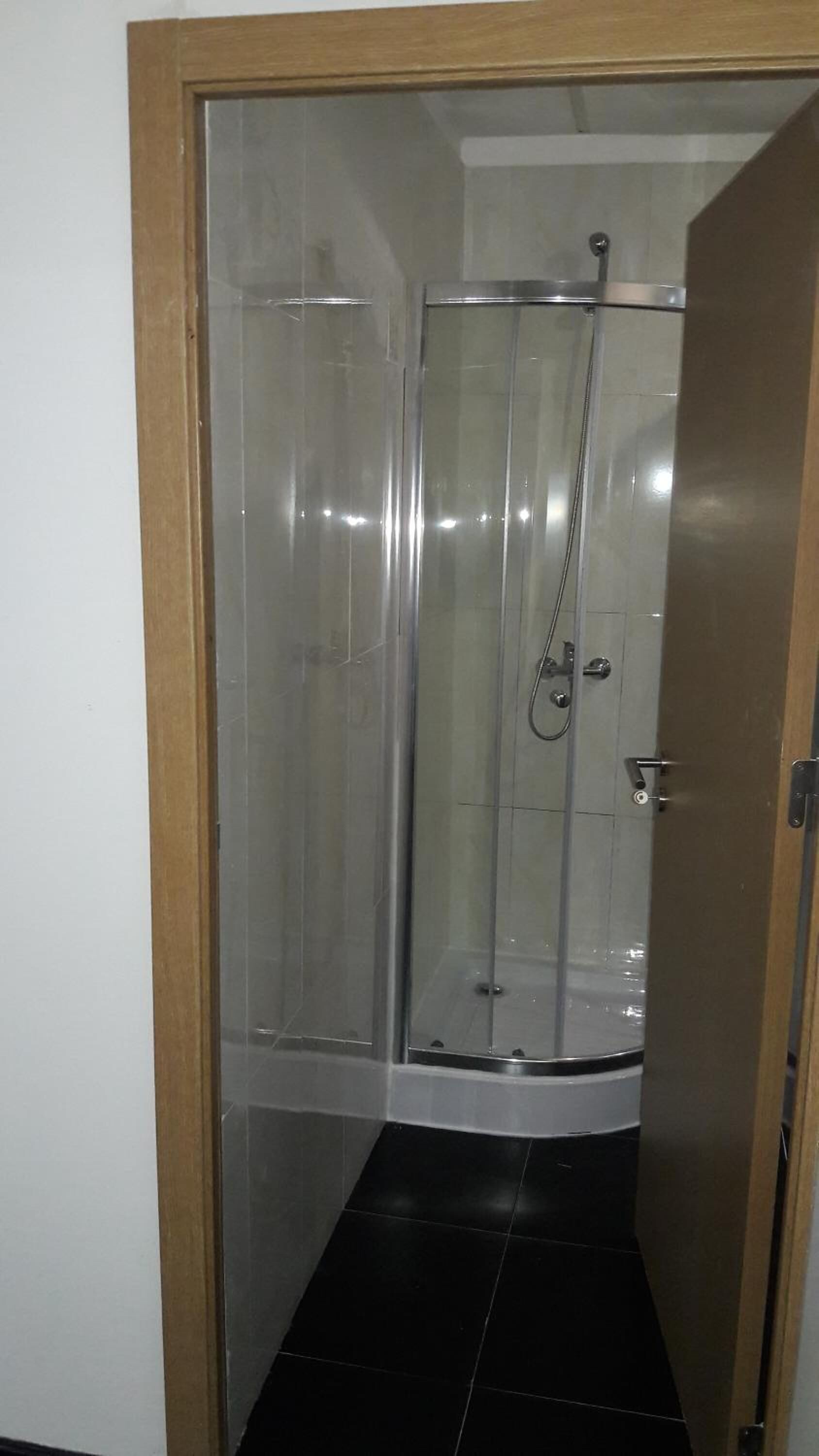 Cabina de ducha con paredes de vidrio, puerta, suelo de baldosas y marco de madera alrededor de la puerta.