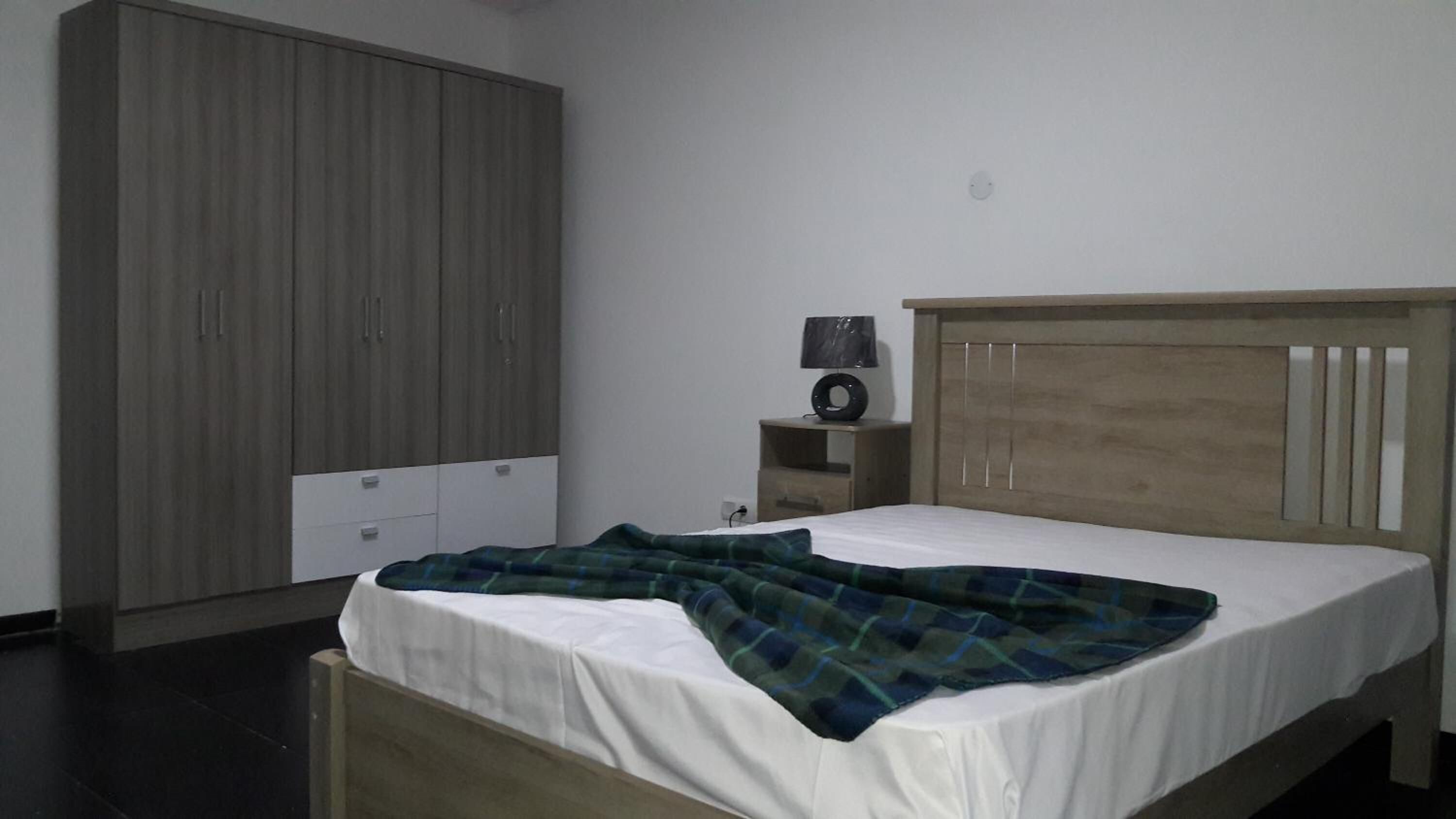 Ein Schlafzimmer mit einem Bett, einem Kleiderschrank, einem Nachttisch mit Lampe und einer Wandsteckdose.
