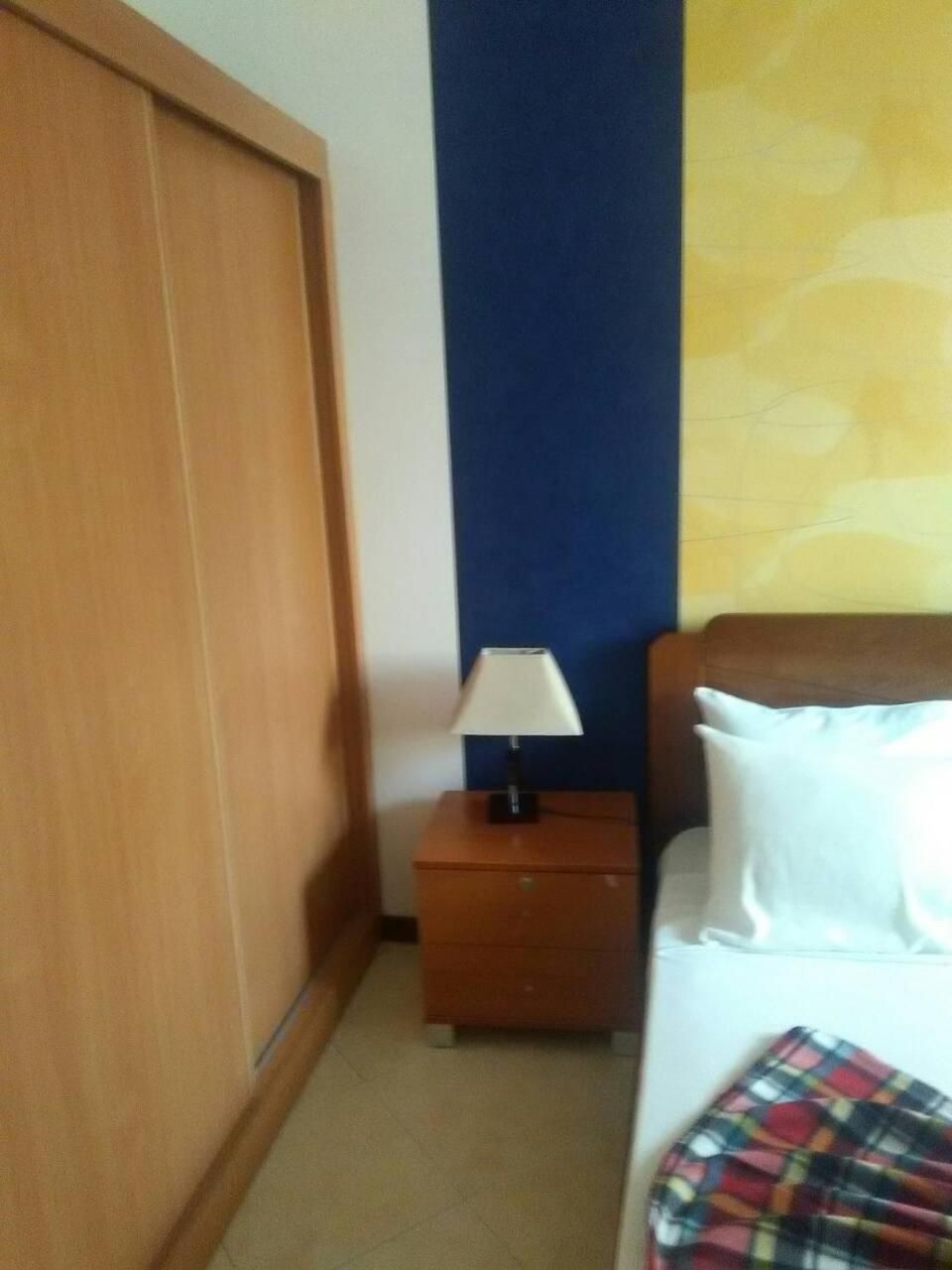 Une chambre avec une armoire en bois, une table de chevet avec une lampe, un lit avec une literie blanche et une couverture colorée.
